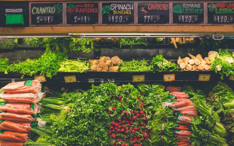 Dans une épicerie, des rangées de légumes frais sont disposées sous des affiches colorées indiquant les prix.