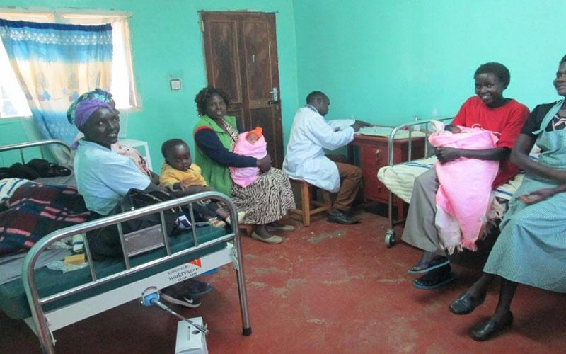 Un groupe de quatre femmes, leurs bébés dans les bras, sourient, assises dans un cabinet médical, tandis qu’un professionnel de la santé travaille à son bureau.
