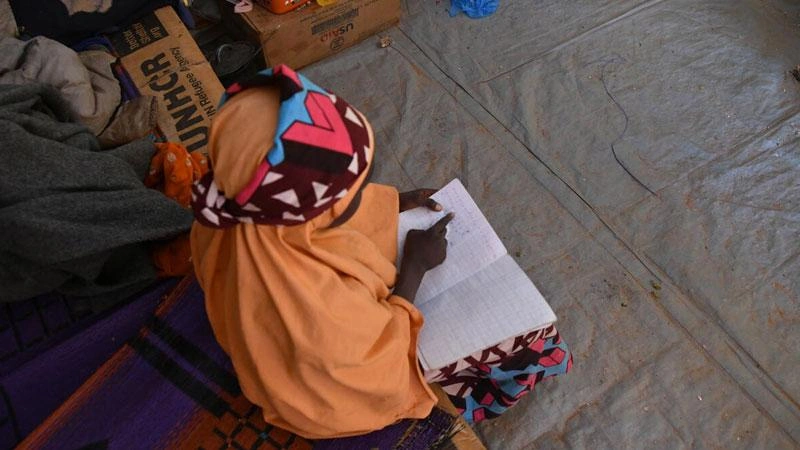Une jeune fille est assise sur un banc dans sa tente, en train de lire un cahier.