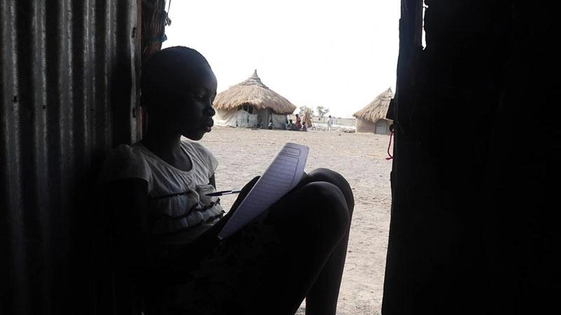 Nyangieth est assise sur le seuil de sa maison, en train de remplir une feuille de travail, avec les huttes de ses voisins en arrière-plan.