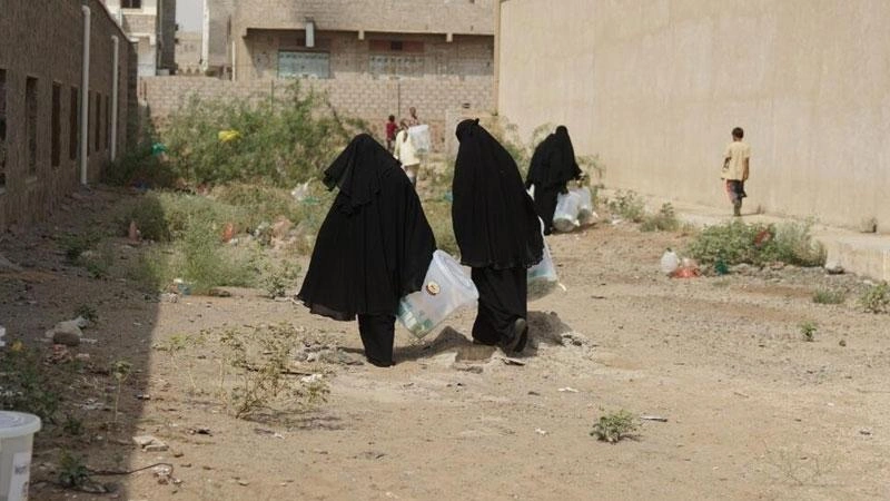Trois femmes portant le niqab traversent un espace désert entre deux immeubles, les bras chargés de sacs contenant des trousses d'hygiène.