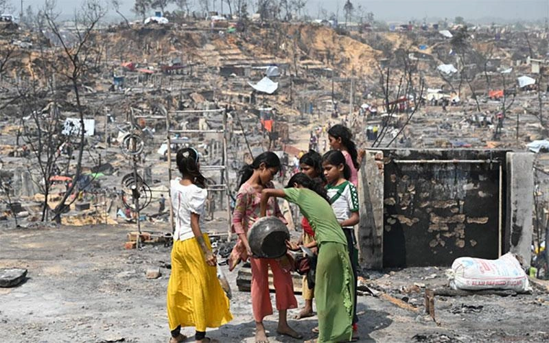 Dans le camp de réfugiés de Cox’s Bazar, un groupe de filles tient un bol ou une casserole et regarde à l’intérieur. Autour d’elles gisent les décombres calcinés du camp.