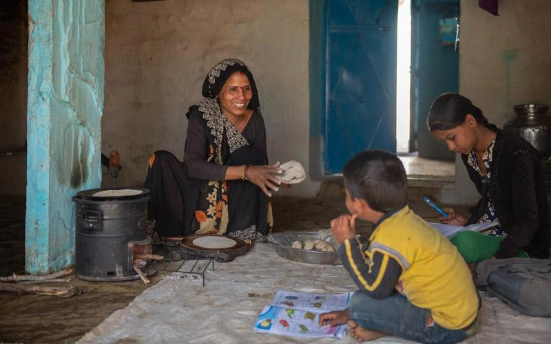 Une femme indienne sourit à deux enfants. Ceux-ci sont assis par terre à l'intérieur, près d'un petit poêle.