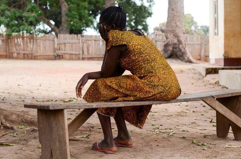 Une femme du Soudan du Sud, vêtue d’une robe jaune, est assise sur un banc de bois, dos à la caméra.