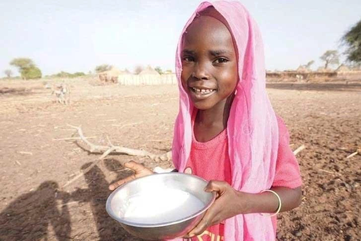 Au Soudan, une petite fille coiffée d’un foulard rose se tient debout au milieu d’un paysage aride, tenant dans ses mains un bol de lait de chèvre frais et blanc. Elle sourit.