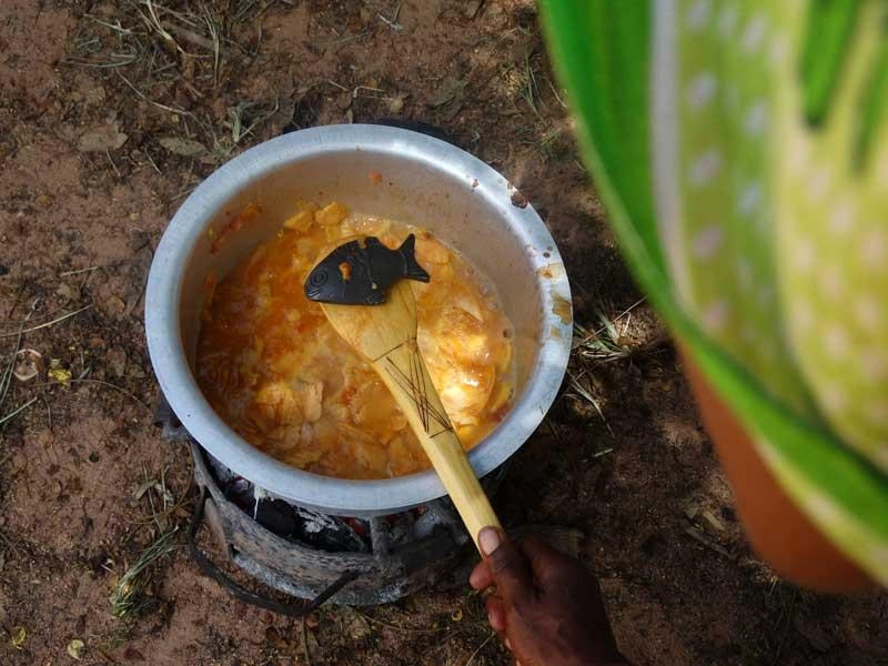 Une femme tient une cuillère en bois posée sur un poisson en fer, au-dessus d’une casserole posée sur un plancher de terre battue.