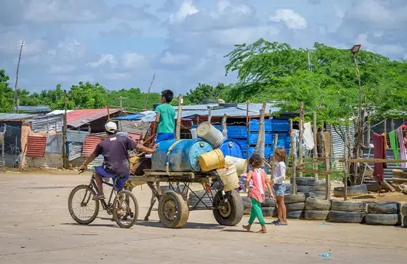 A child rides on top of a cart carrying buckets, while two children walk at the back and another rides their bicycle beside it. (Colombia, 2023)
