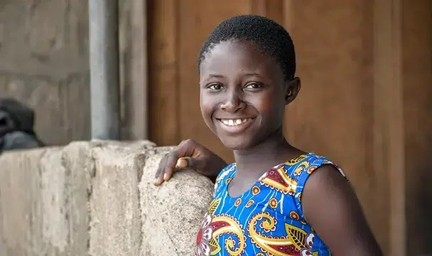 A young girl in a blue patterned shirt, smiles. (Ghana, 2024)