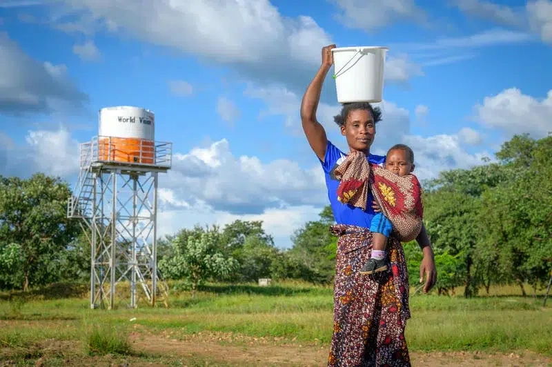 Une mère tenant son bébé dans les bras va chercher de l’eau potable, un réservoir d’eau de World Vision se détachant en arrière-plan.