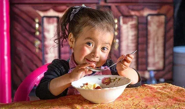 A little girl smiles at the camera while holding a spoon in their hand.