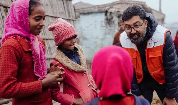 A man wearing a World Vision vest looks down and talks to three children outside their home in Nepal.