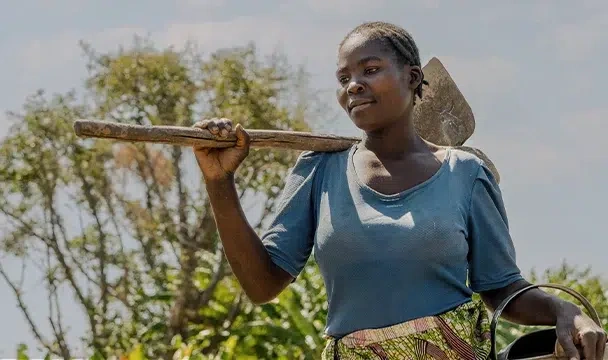 A woman in a blue shirt carrying a hoe and watering can outdoors, surrounded by greenery.