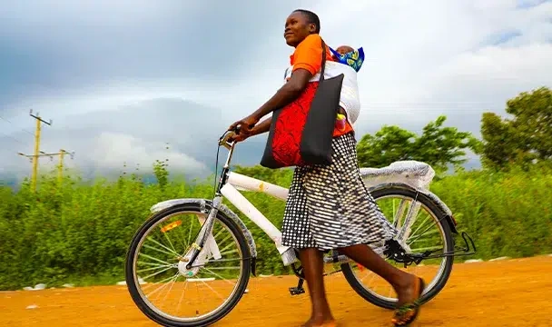 A woman carrying a child on her back tries out her bicycle after receiving it.