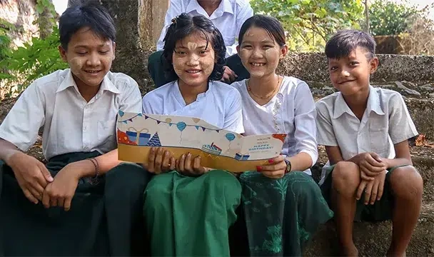 Children in school uniforms sit outdoors, reading a colorful booklet together.