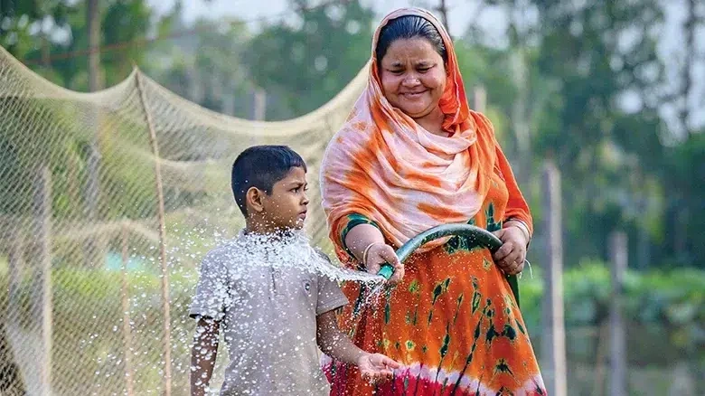 A woman wearing an orange dress waters her vegetables with a young boy beside her. (Bangladesh, 2025)