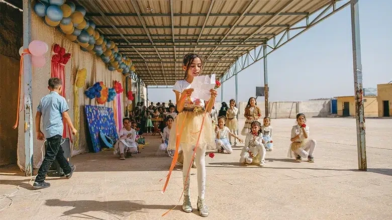 A young girl wearing a white shirt and yellow skirt is reading a letter after participating in a community event. (Syria, 2024)