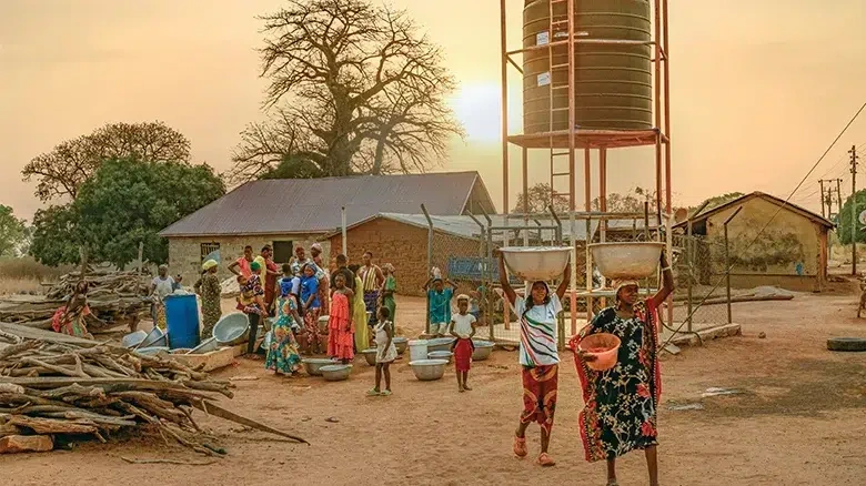 An enormous water tank towers over a group of houses as women, men and children gather around to fill large metal bowls and buckets with clean water.