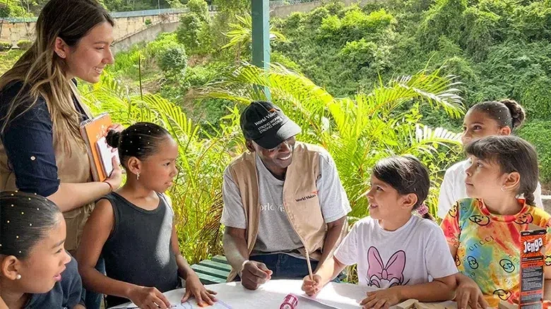 World Vision staff leading outdoor educational activity with children, featuring drawing materials and games on a table in a lush, green setting.