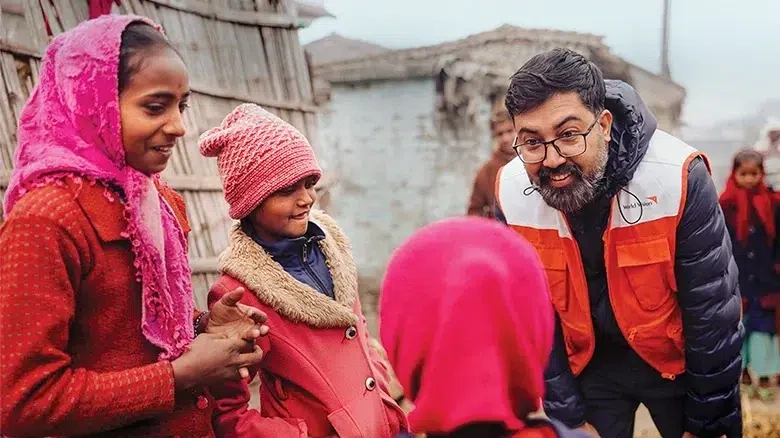 A man wearing a World Vision vest looks down and talks to three children outside their home in Nepal.