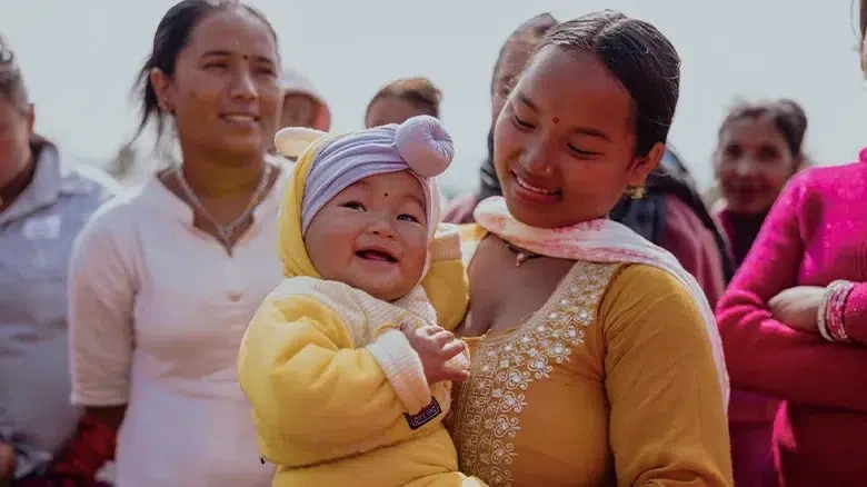 Mother holding her baby in a yellow outfit while standing among a group of women during a community gathering.
