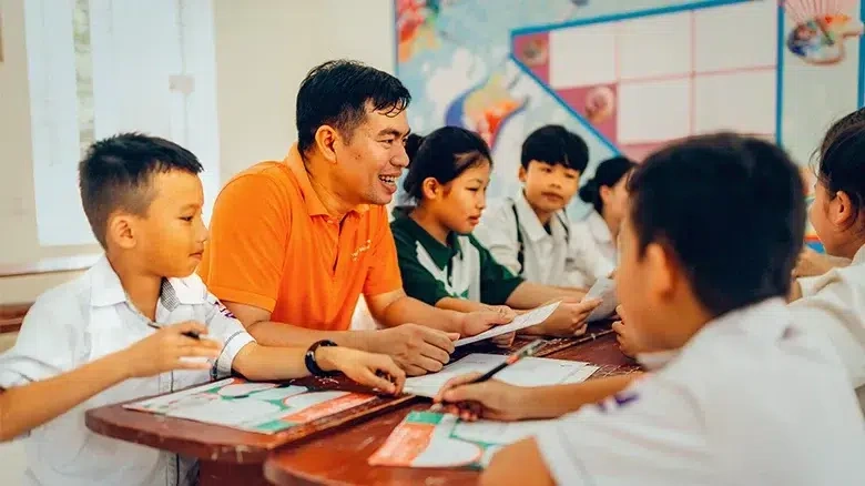 A male teacher wearing an orange t-shirt is sitting with his young students as they participate in a classroom activity. (Vietnam, 2024)