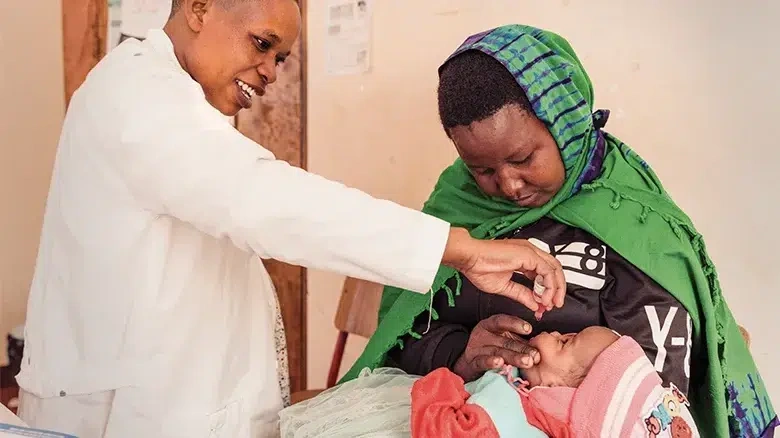 A woman wearing a green headscarf holds a baby as a healthcare worker examines her. (Tanzania, 2025)
