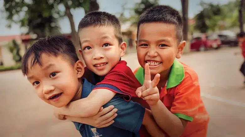 A little boy wearing a red shirt clings to an older boy’s back as another boy in an orange shirt makes a peace sign behind them.