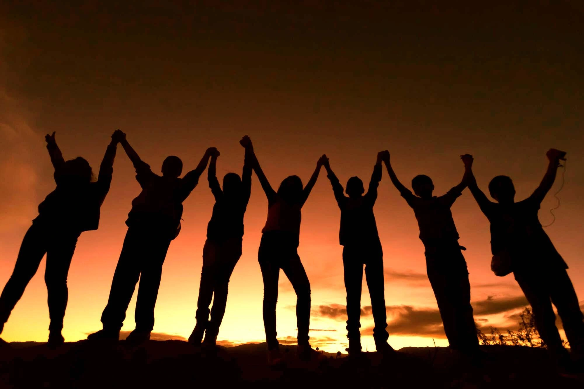 Children stand raising their arms in a silhouette against an orange sunset.