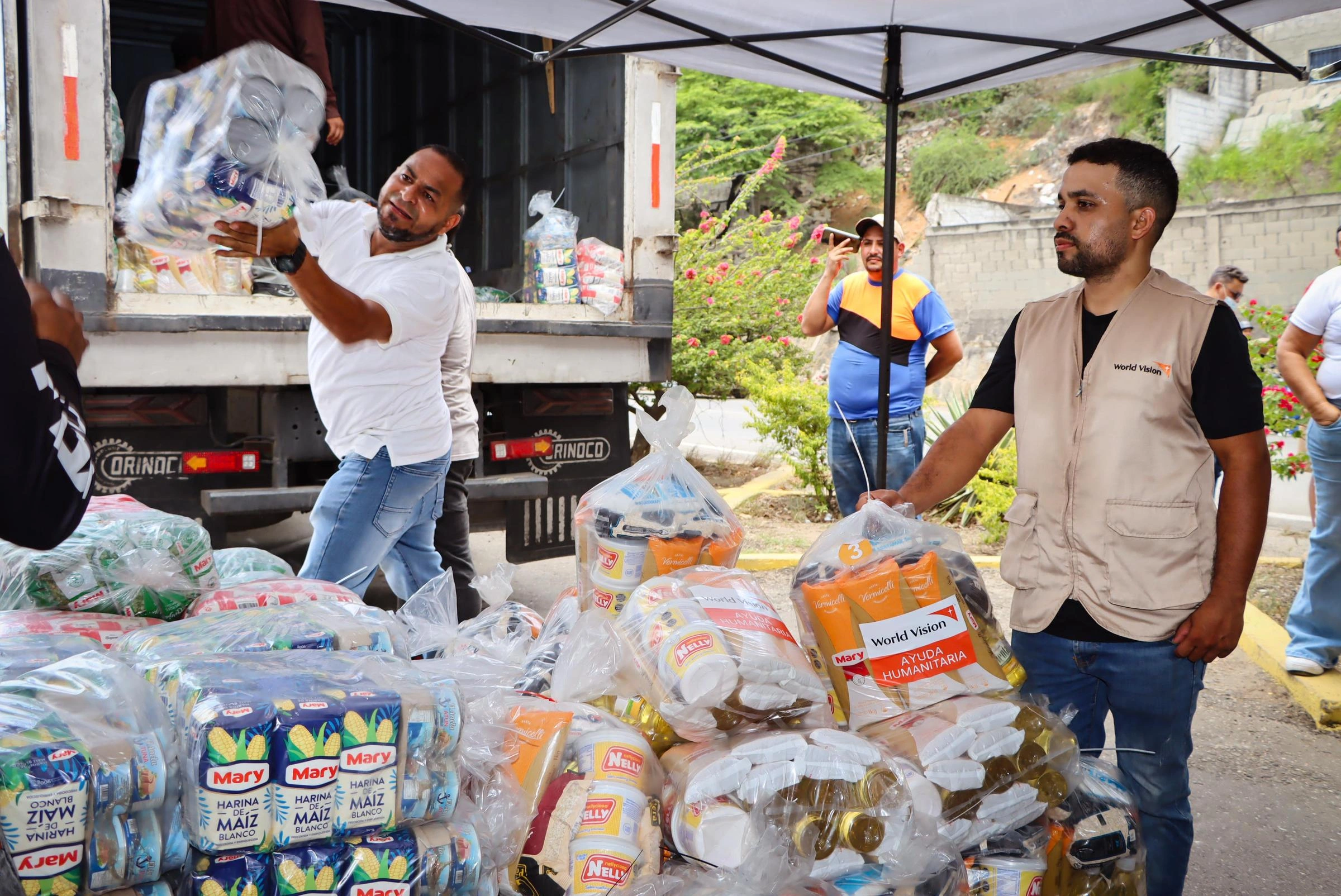 A volunteer in a World Vision vest watches as another person passes a large bag of food supplies.