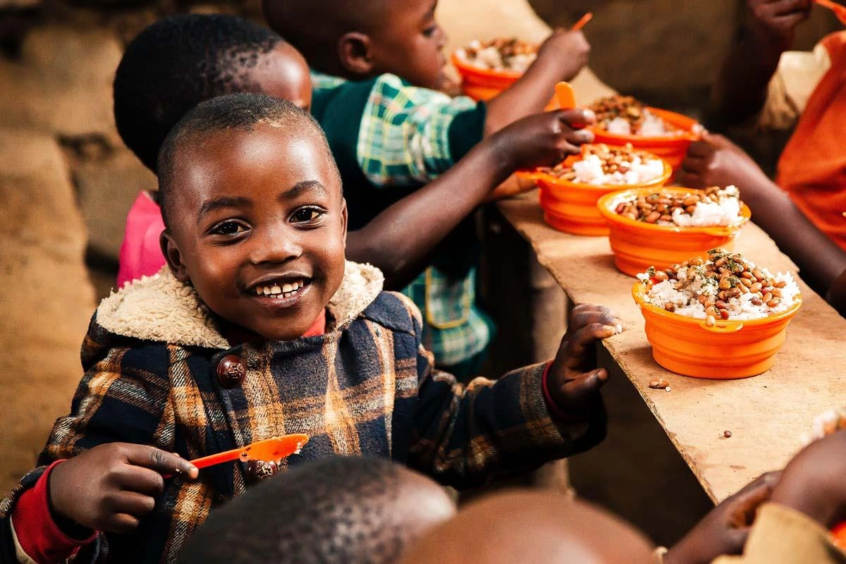 A child smiles facing the camera, while other children eat from their orange bowl and spoon sets provided by World Vision.