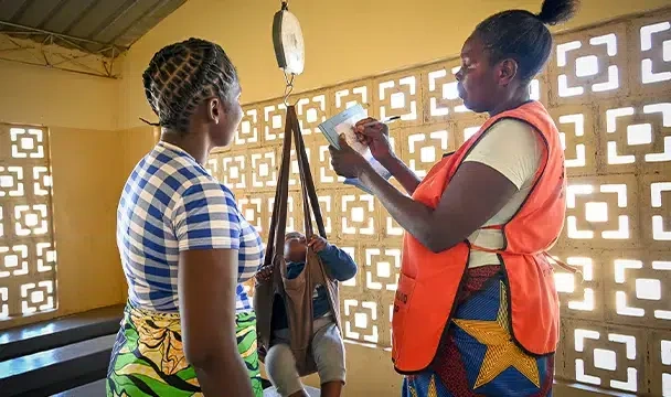 Medical professional weighs infant and writes the weight on paper as the mother stands beside her.