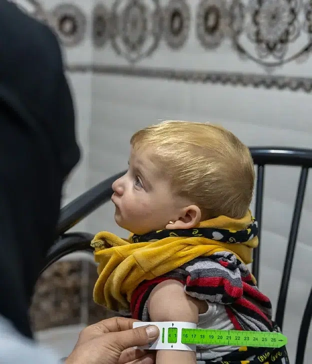 A young child in a yellow sweater has their arm measured at a health clinic.