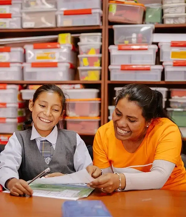 A girl in a school uniform sits beside a world vision staff at a table looking at a book.