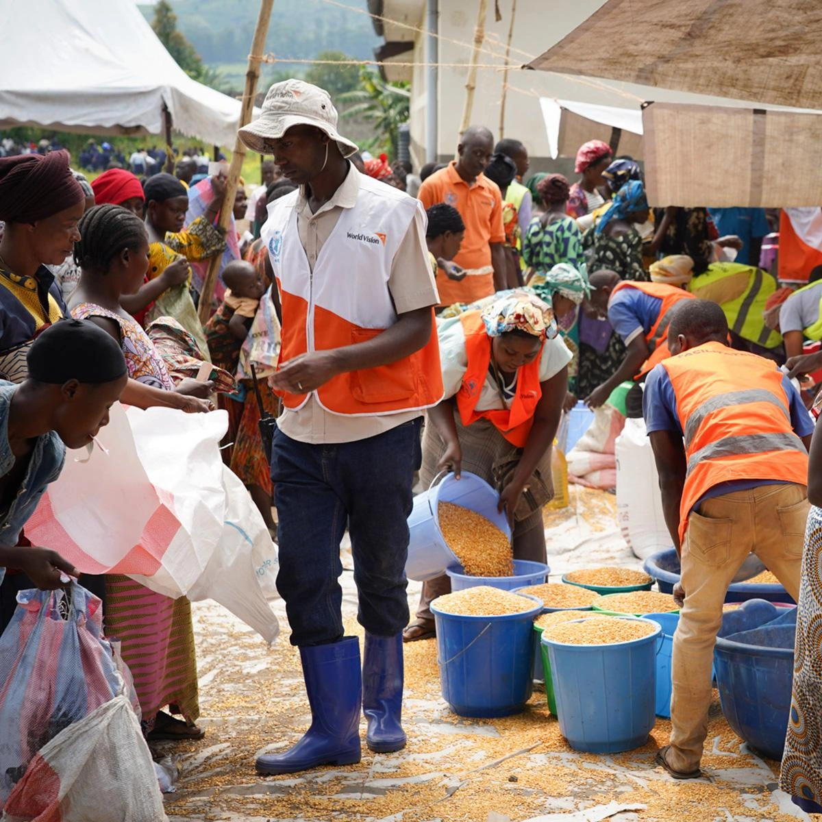 Relief workers in orange safety vests distribute food to community members, scooping grain into buckets and bags at an outdoor aid distribution site.