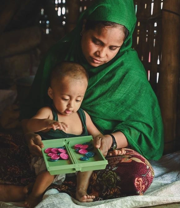In Bangladesh, a woman in a vibrant green shawl sits on the ground with an infant playing in her lap.