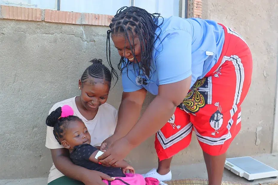 A healthcare worker measures a young girl’s arm to monitor growth while she sits in her mother’s lap.