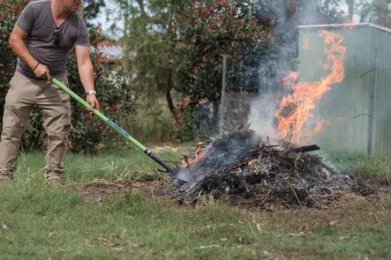 Man burning brush in a backyard. He is holding a poker as he tends the fire.