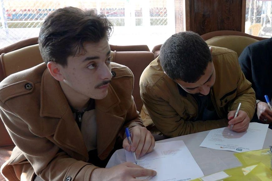 Two young men write on papers on a table, as one looks up and off-camera.