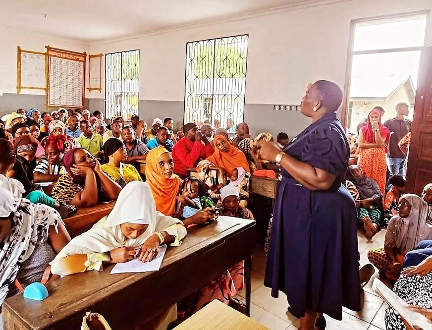 A woman in a navy dress speaks to a crowded roomful of students who are seated at desks and on the floor.