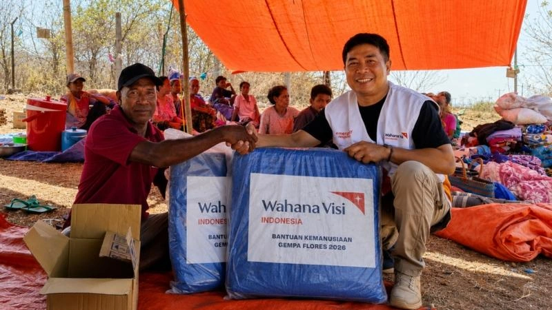 A World Vision staff member hands a package of emergency shelter supplies to a community member under a tarpaulin shelter, while women and children sit together behind them.