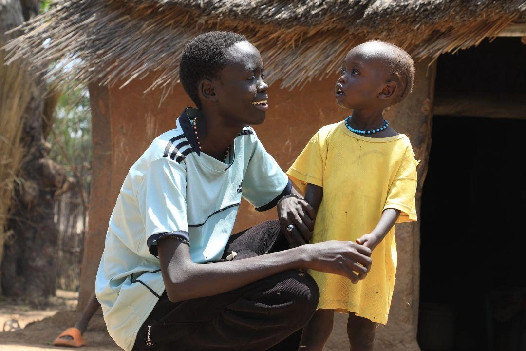 A teenage boy sits next to his little sister outside.