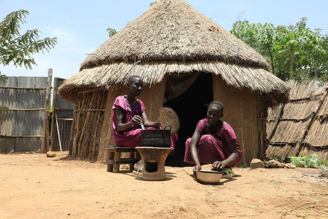 A grandmother and granddaughter prepare food outside their home that has a grass roof.