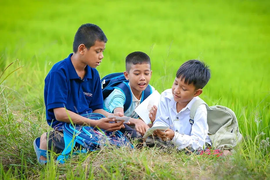 Three boys sit together on a grassy field, looking at a photograph and discussing it with one another.