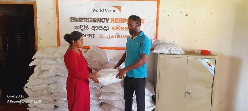 An aid worker hands relief supplies to a woman beside stacked sacks under a World Vision emergency response banner in Karachchi, Sri Lanka.
