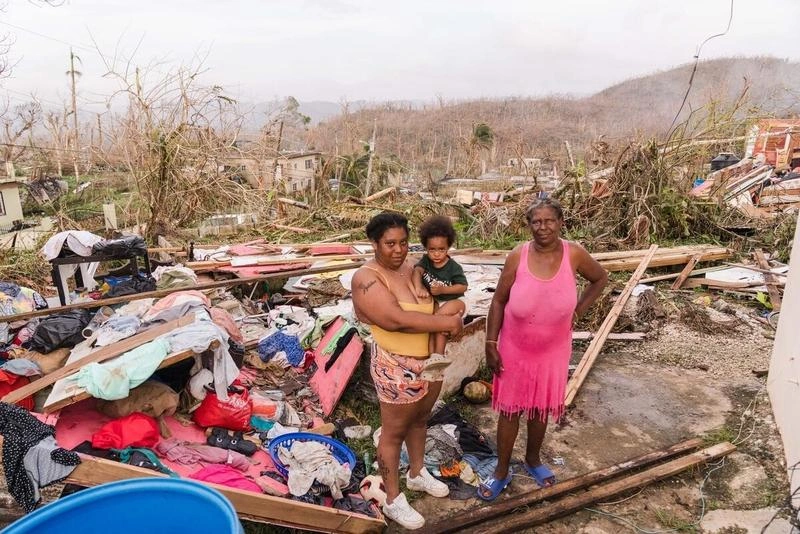 Two women and a young child stand among scattered clothing, timber and household debris outside a storm-damaged home in Chester Castle, Jamaica.