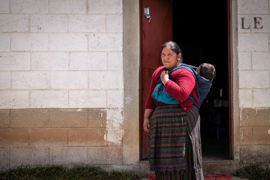 A woman stands in front of a building built from concrete blocks, a sleeping baby on her back.