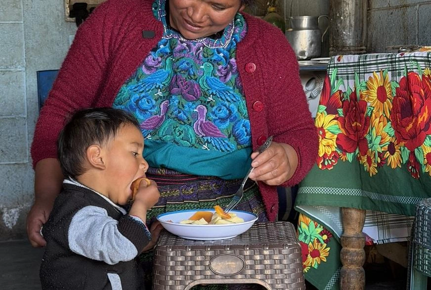 A toddler feeds himself food from a bowl while a woman assists with a fork.