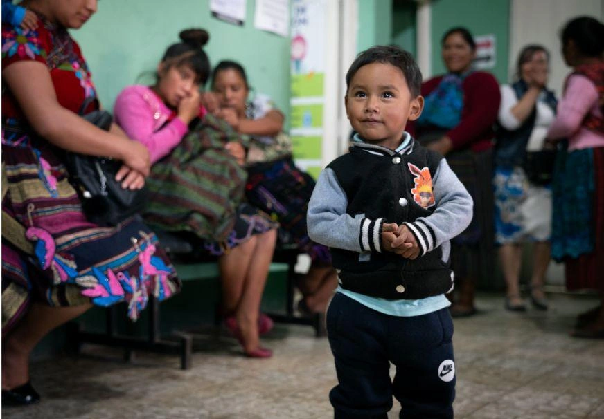 A toddler stands in a waiting room full of people, smiling to someone off camera.
