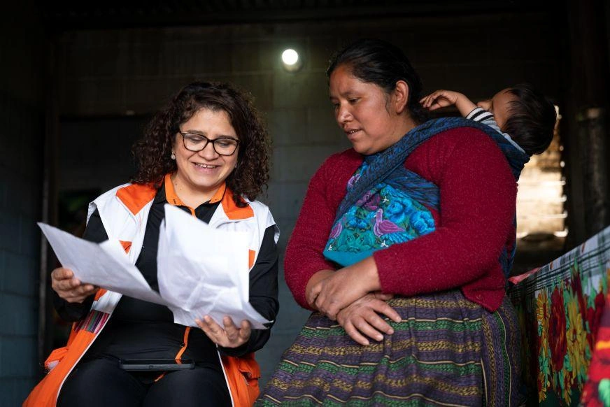 Two women sit together, one holding papers and wearing a World Vision vest. The second woman carriers a toddler on her back.