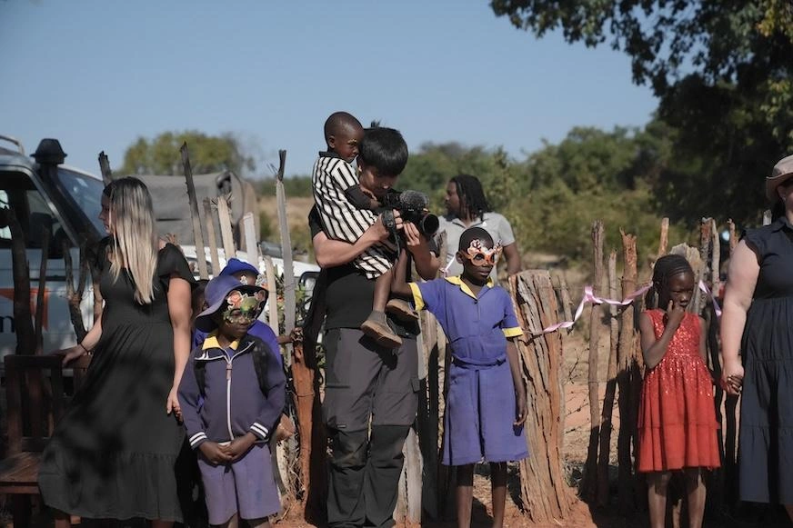 A man wearing black balances a young child in his arms as he looks through a camera. Other children look off camera to see what he is filming.